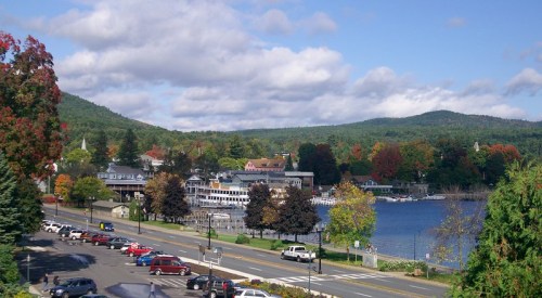 A scenic view of a lakeside town with colorful trees, boats, and mountains under a partly cloudy sky.