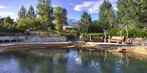 A serene natural hot spring surrounded by trees and benches, with mountains in the background under a clear blue sky.