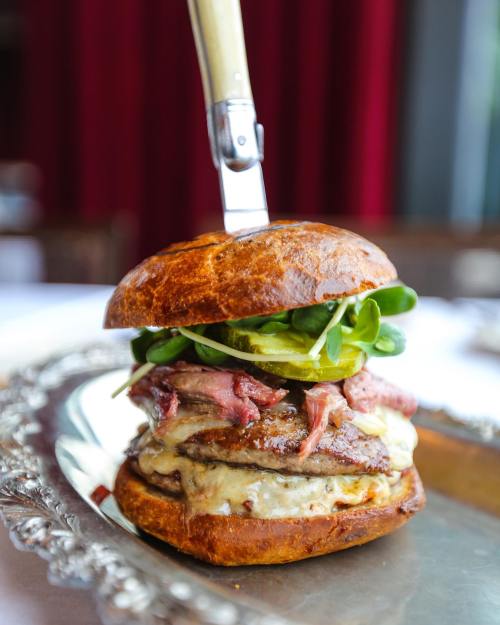 A gourmet burger stacked with meat, cheese, greens, and pickles, served on a silver platter with a knife inserted.