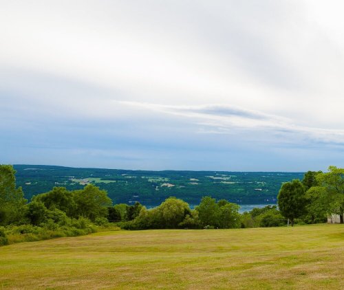 A serene landscape featuring green fields, trees, and a distant view of rolling hills under a cloudy sky.