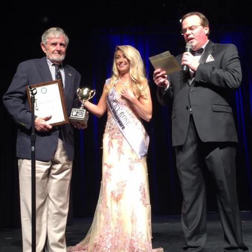 A man receives an award from a woman in a gown, while another man reads from a paper on stage.