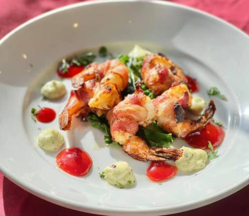 A plate of bacon-wrapped shrimp on a bed of greens, garnished with sauces and herbs.