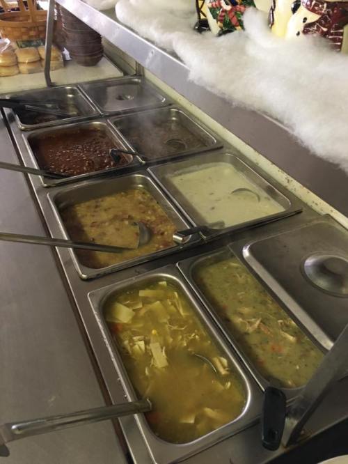 A buffet-style setup with several metal trays containing various soups and stews, steam rising from some.