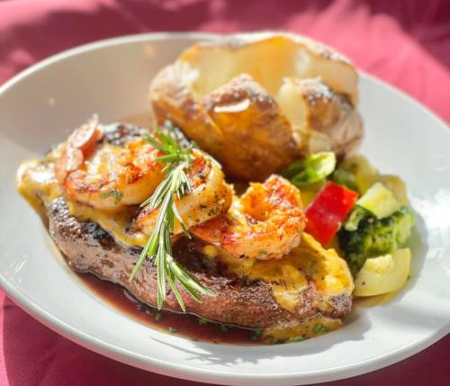 A plate featuring grilled shrimp on a steak, a baked potato, and mixed vegetables, all garnished with herbs.