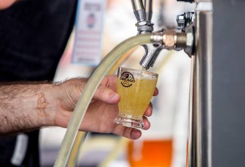 A hand pours a light-colored beverage into a small glass at a tap, with a logo visible on the glass.