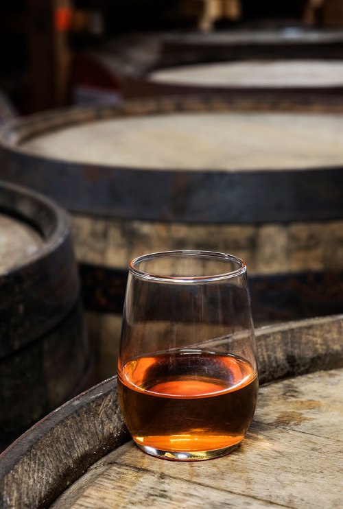 A glass of amber liquid sits on a wooden barrel, with more barrels blurred in the background.