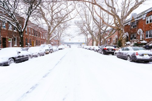 Snow-covered street lined with houses and parked cars, with bare trees and a cloudy sky above.