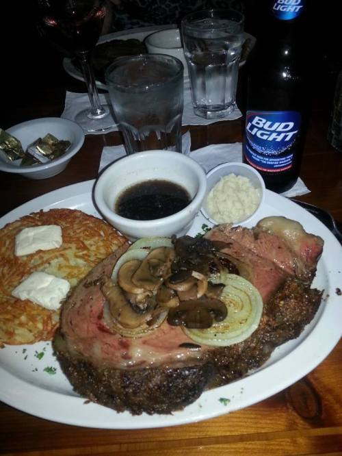 A plate of prime rib with mushrooms and onions, hash browns, a side of sauce, and a Bud Light beer.
