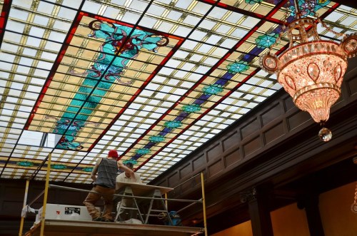 Two workers on scaffolding install or repair a colorful stained glass ceiling, with an ornate chandelier nearby.