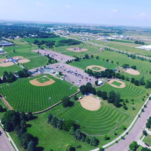 Aerial view of multiple baseball fields surrounded by green grass and trees, with a clear blue sky above.