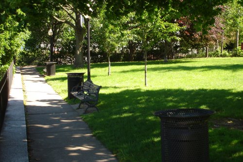 A sunny park scene featuring a grassy area, benches, and trash cans along a paved walkway.