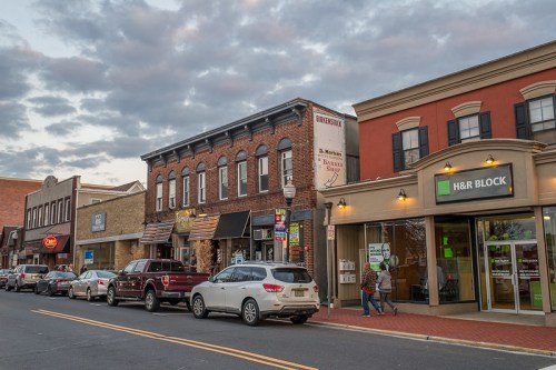 A street view of a small town with shops, parked cars, and a cloudy sky in the background.