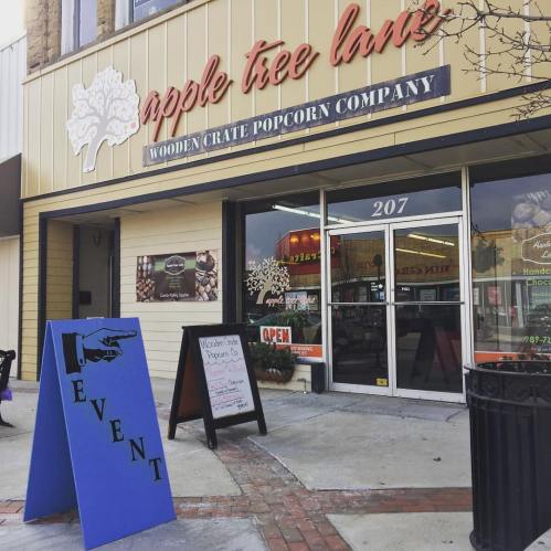 Storefront of Apple Tree Lane with a blue event sign and a chalkboard menu outside the Wooden Crate Popcorn Company.