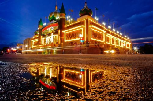 A brightly lit building with green and gold accents reflects in a puddle at dusk, surrounded by a darkening sky.
