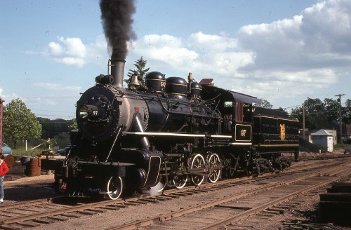 A vintage steam locomotive with black and gold detailing, emitting smoke, parked on railway tracks under a blue sky.