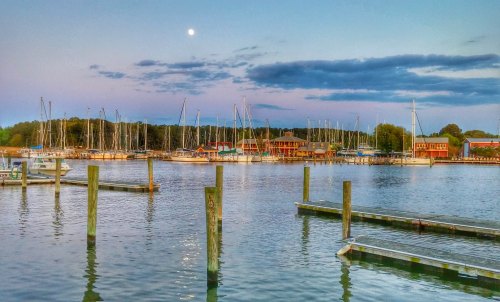 A serene marina at dusk, featuring sailboats, a calm water surface, and a full moon in a clear sky.