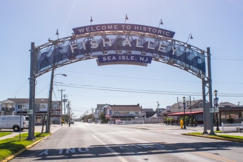 Archway sign reading "Welcome to Historic Fish Alley, Sea Isle, NJ" over a sunny street with buildings in the background.