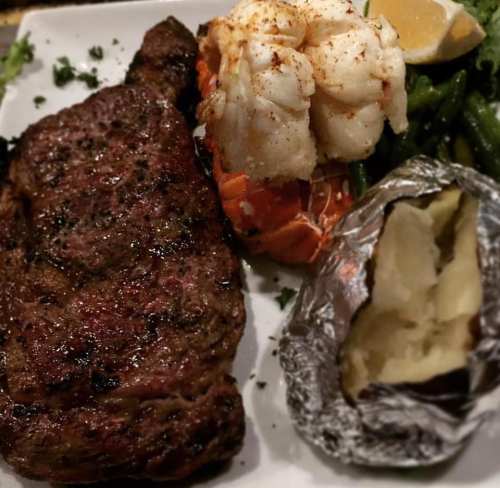A plate featuring a grilled steak, lobster tail, green beans, and a baked potato wrapped in foil.