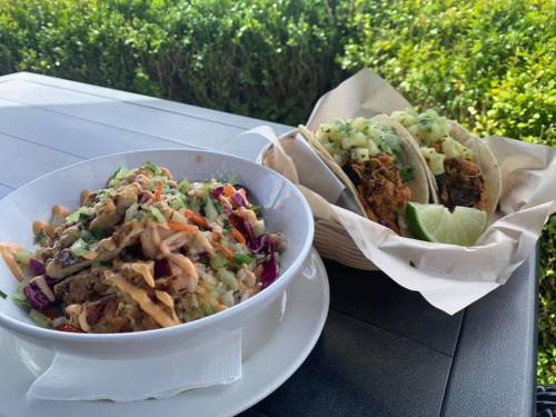 A bowl of colorful slaw with meat beside two tacos garnished with cilantro and lime wedges on a table.