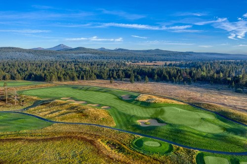 Aerial view of a golf course surrounded by lush greenery and mountains under a clear blue sky.