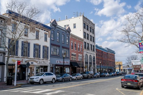 A street view featuring historic buildings, shops, and parked cars under a partly cloudy sky.