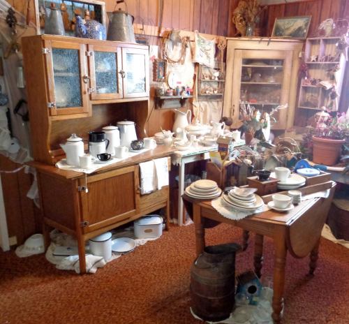 A cozy vintage kitchen with wooden furniture, dishes, and decorative items arranged on shelves and tables.