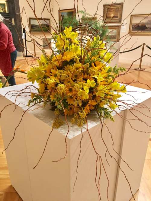 A vibrant floral arrangement featuring yellow flowers and twigs, displayed on a white pedestal in an art gallery.