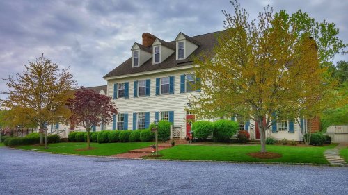 A large, two-story white house with blue shutters, surrounded by green trees and a gravel driveway.