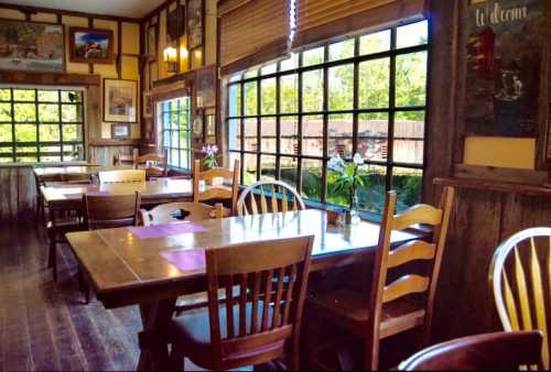 Cozy restaurant interior with wooden tables and chairs, large windows, and a view of greenery outside.