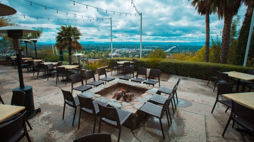 Outdoor seating area with a fire pit, surrounded by palm trees and a view of the city under a cloudy sky.