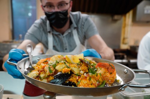 A person in a mask and gloves holds a large dish of seafood paella, garnished with lemon and herbs.