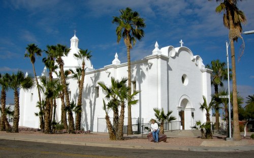 A white church with palm trees in front, set against a blue sky with clouds. A person stands near the entrance.