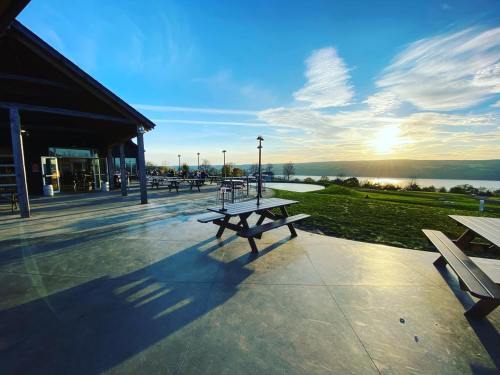 A scenic view of a lakeside area at sunset, featuring picnic tables and a building with large windows.