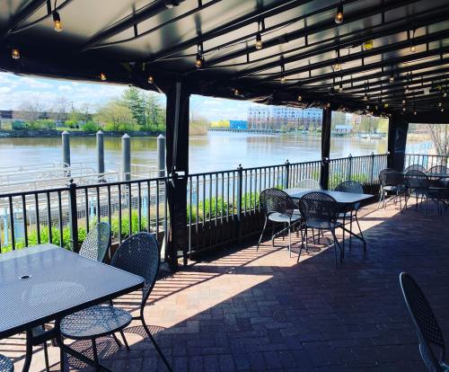 Outdoor seating area with black tables and chairs overlooking a river, under a canopy with string lights.