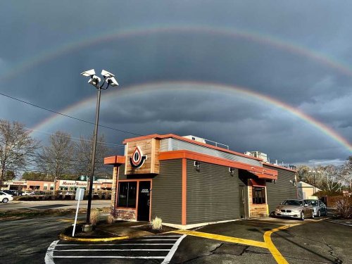 A building with a logo under a double rainbow against a cloudy sky, with a parking lot and nearby trees.