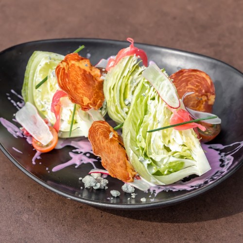 A fresh salad featuring lettuce wedges, crispy toppings, pickled onions, and a colorful dressing on a black plate.