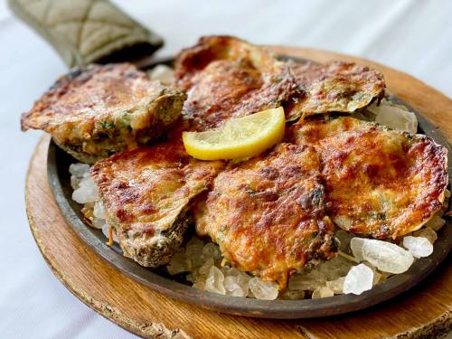 Grilled oysters topped with cheese and herbs, served on a sizzling platter with a lemon wedge.