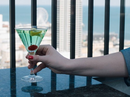 A hand holds a green cocktail with a cherry and lemon slice, set against a city skyline and ocean view.