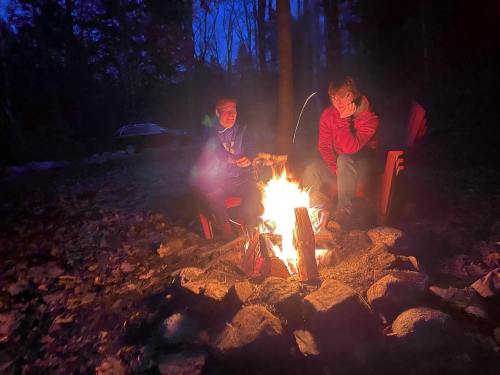 Two people sit by a campfire in a wooded area at dusk, surrounded by rocks and fallen leaves.