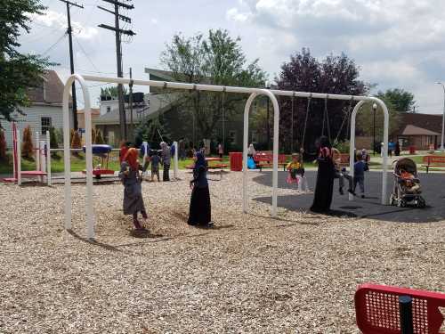 A playground scene with children and adults on swings, surrounded by trees and play equipment on a sunny day.