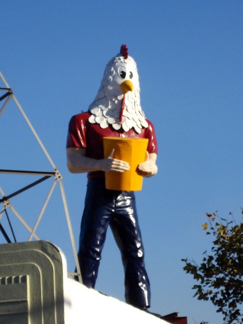 A large statue of a person with a chicken head, holding a yellow bucket, stands against a clear blue sky.