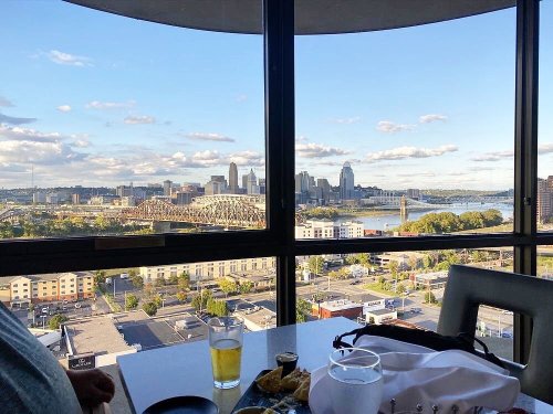 A city skyline view from a high-rise window, featuring a river, bridge, and a table with drinks and a bag.
