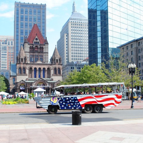 A decorated bus with an American flag design parked in a city square, surrounded by tall buildings and a historic church.