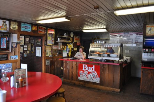 A cozy diner interior with a red table, wooden counter, and vintage signs, featuring a staff member at the grill.