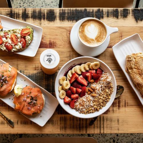 A wooden table with a breakfast spread: avocado toast, smoked salmon, a smoothie bowl, and a latte.