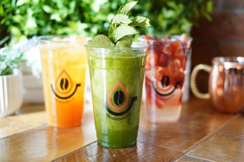 Three colorful drinks in clear cups on a wooden table, featuring a green mint drink in the foreground.