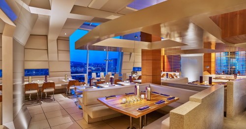 Modern restaurant interior with elegant seating, wooden tables, and large windows showcasing a city view at dusk.