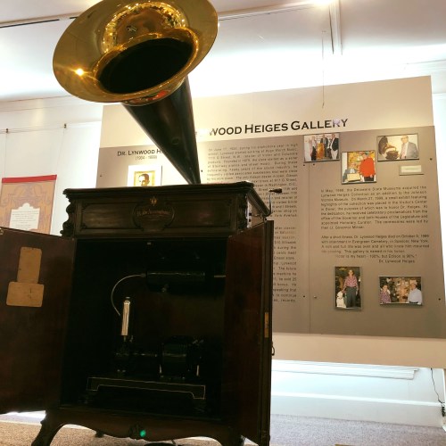A vintage gramophone displayed in a gallery, with an informational sign about Dr. Lynwood Heiges in the background.