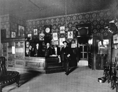 Historic black-and-white photo of a vintage hotel lobby with three people, ornate wallpaper, and wooden furnishings.