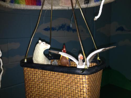 A woven basket with toy animals, including a bear, a rooster, and a duck, suspended from a colorful balloon.